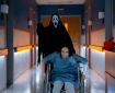 A person in a wheelchair leans forward in a hospital hallway while a tall, black-robed figure wearing a white ghost mask stands behind them.