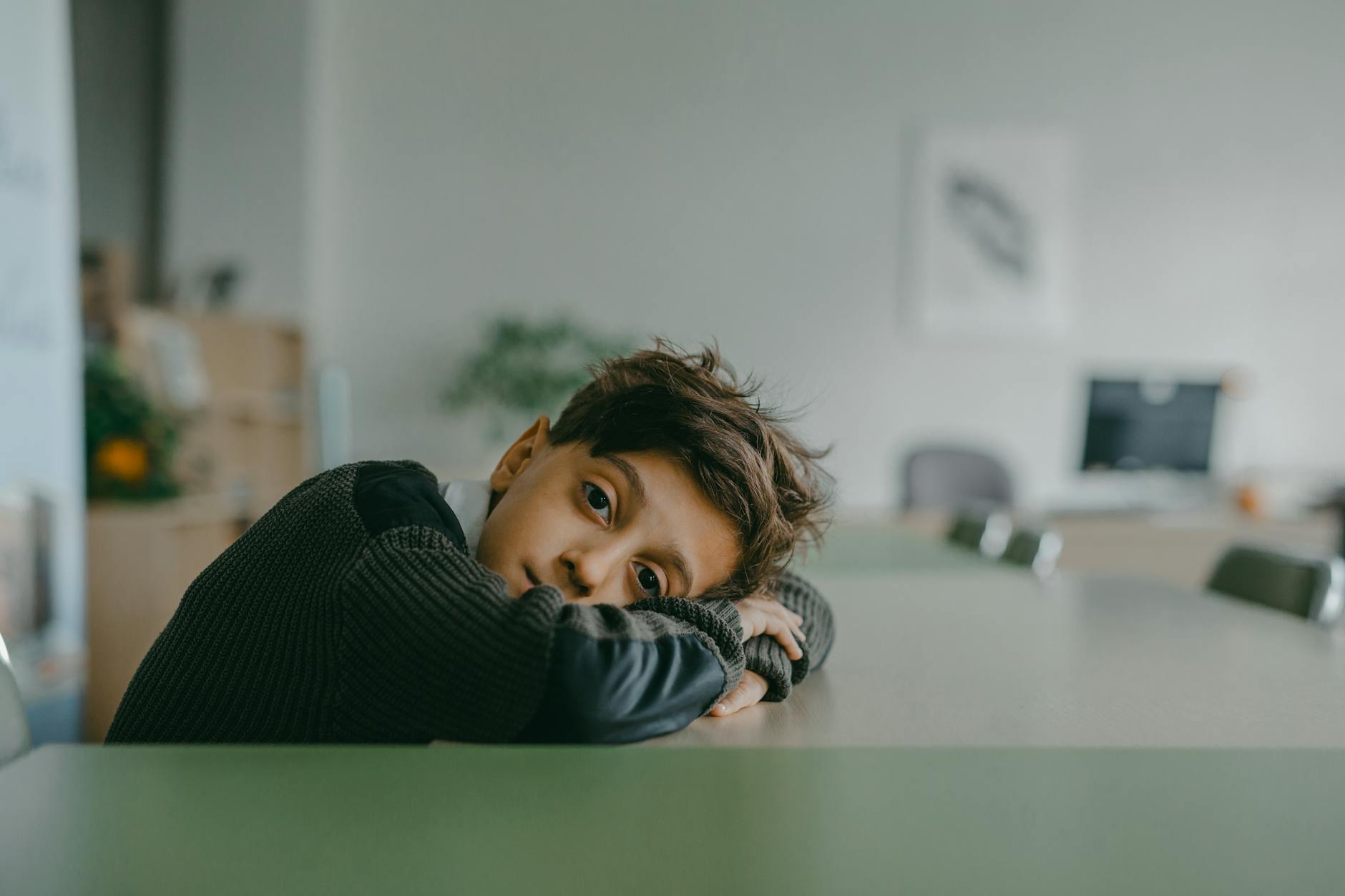 a boy wearing long sleeves sweater resting head on a desk
