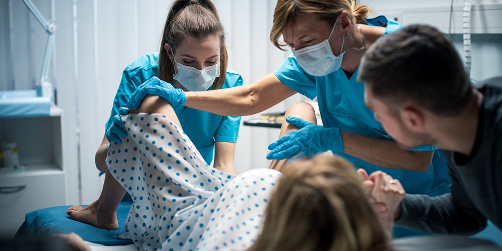 In the Hospital Woman in Labor Pushes to Give Birth, Obstetricians Assisting, Husband Holds Her Hand. Modern Delivery Ward with Professional Midwives. Back View Footage.