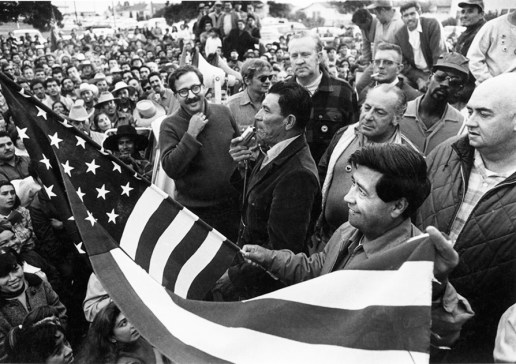 Cesar Chavez waves an American flag to counter Red-baiting during an organizing rally. To his right is Chavez confidant Marshall Ganz (wearing glasses). Directly behind the speaker (in plaid shirt) is Bill Kircher, AFL-CIO Director of Organizing and liaison to the UFW, and members of the Seafarers Union. Salinas, California, 1970. Photo Bob Fitch