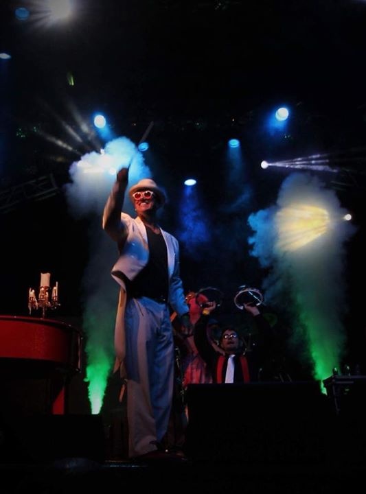 Elton Dan on stage, standing in front of his band, conducting the audience in a sing-along.