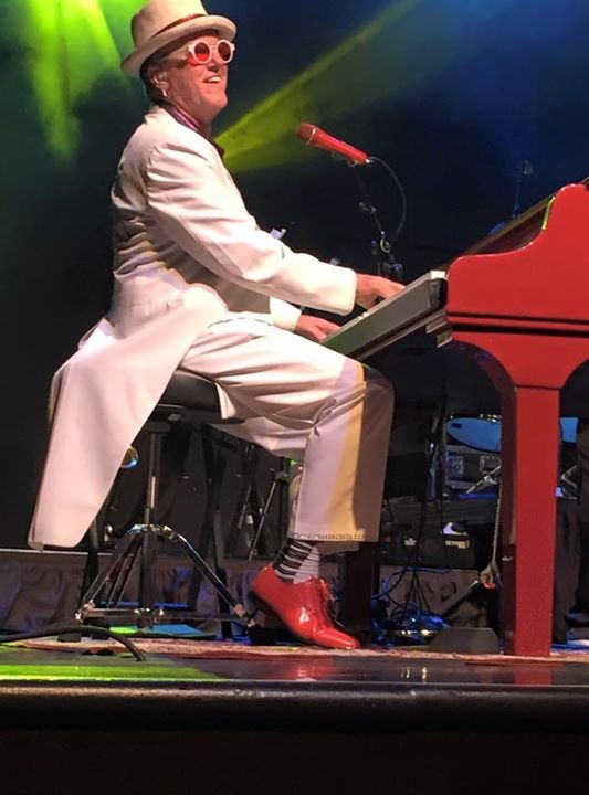 Elton Dan wearing red shoes, piano socks and his white suit with tails. He's on stage, playing piano and smiling at the audience.