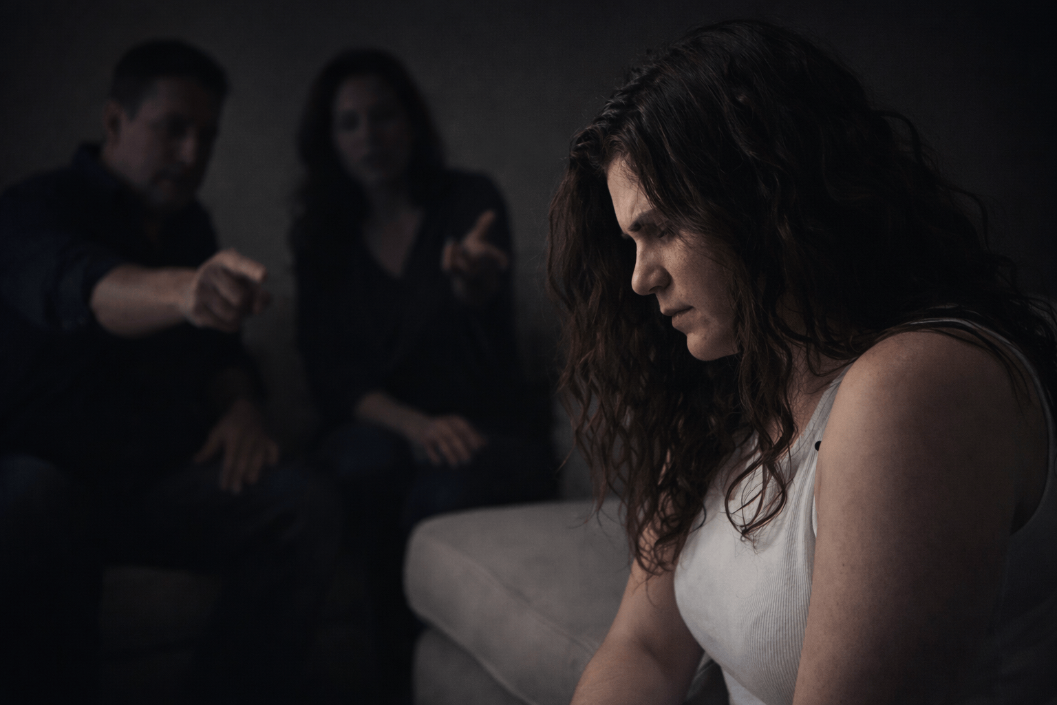 Woman sitting in tense family conversation while two shadowed figures gesture in the background, illustrating emotional unsafety and interrogation dynamics.