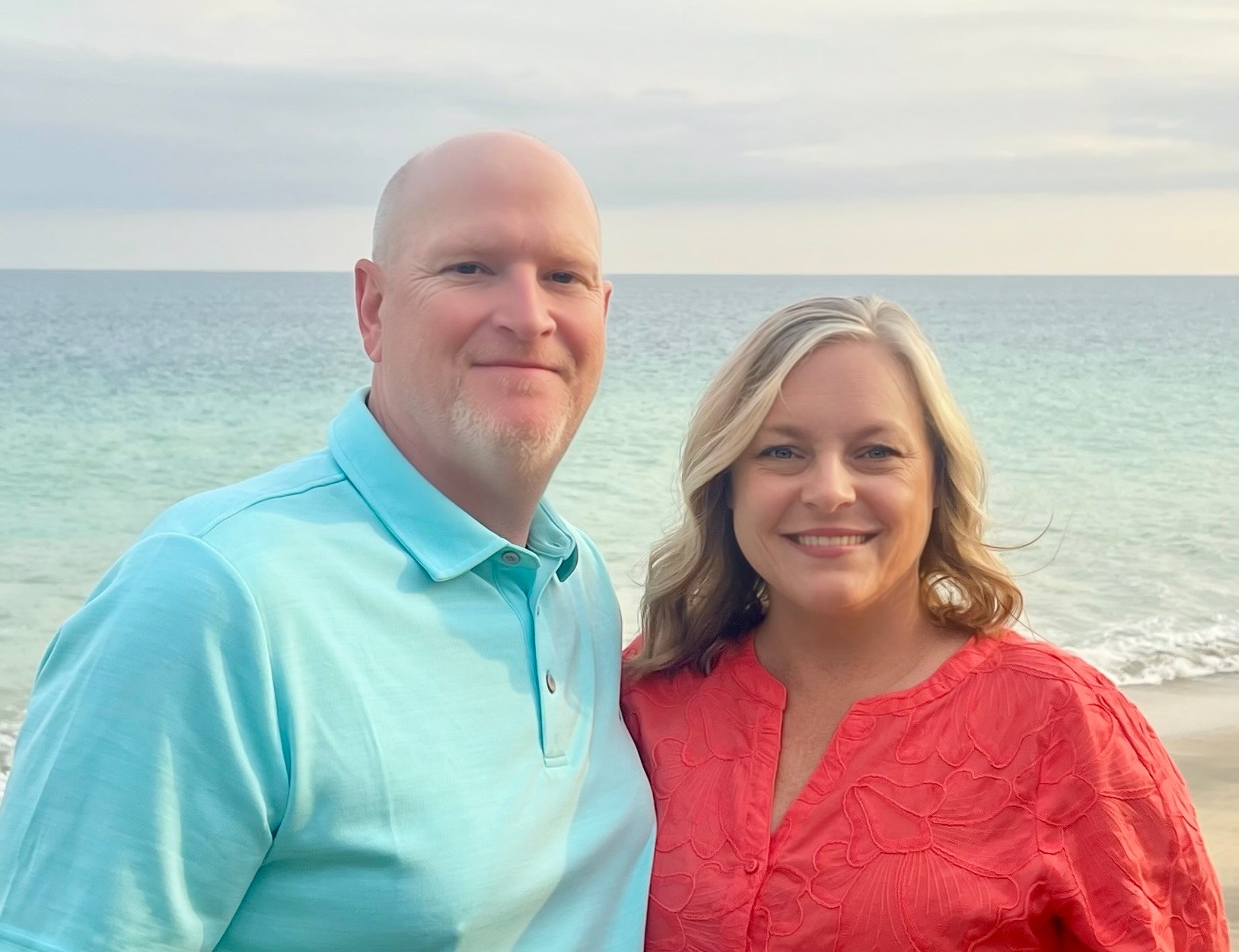 Smiling couple standing together by the ocean, with the woman in a coral top and the man in a turquoise polo