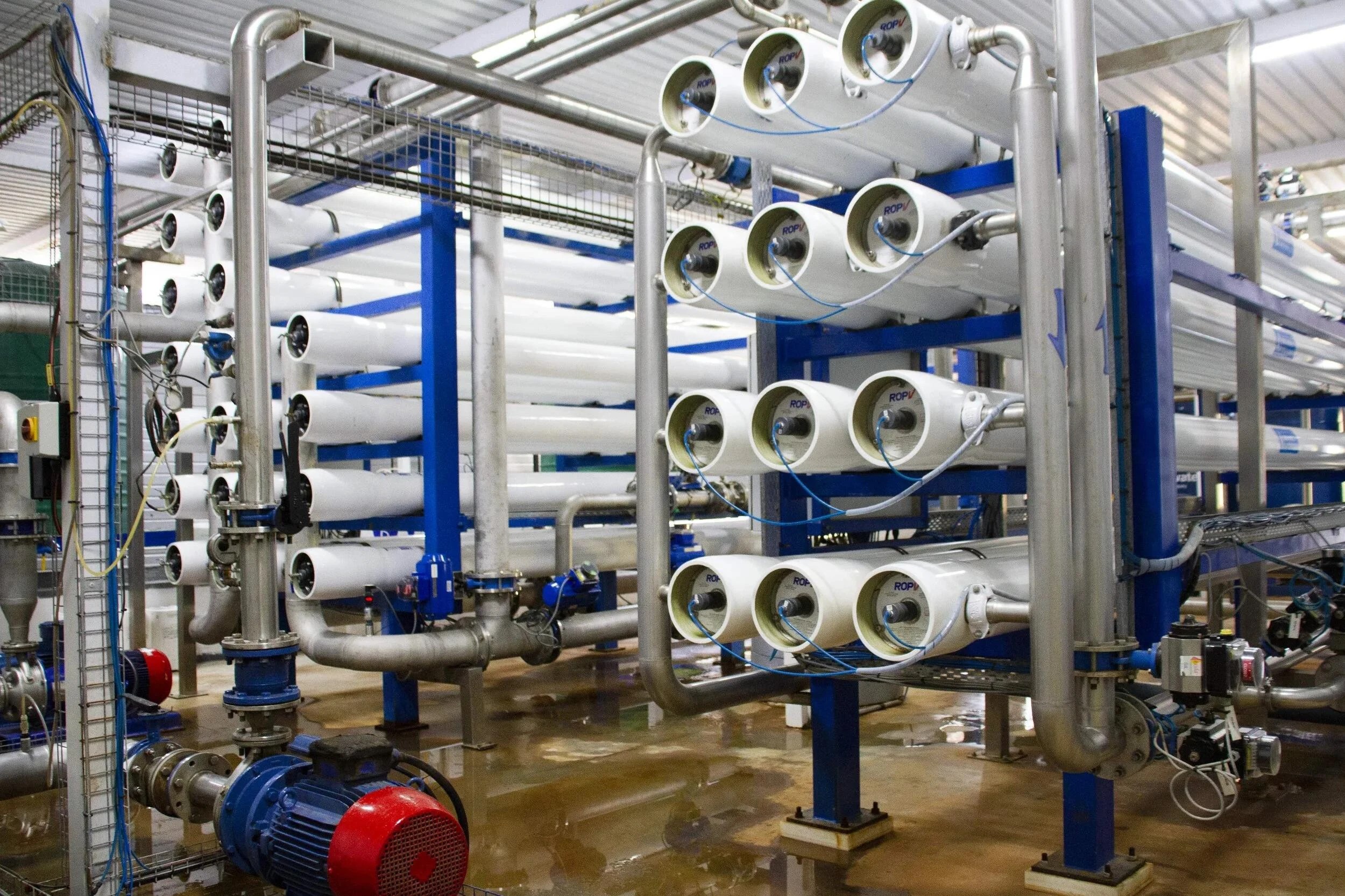 Industrial piping system in a factory: white pipes on blue racks with valves, cables, and a red electric motor in the foreground.