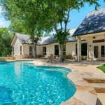 A curved backyard swimming pool with clear blue water next to a white house, stone patio, green lawn, and tall trees.
