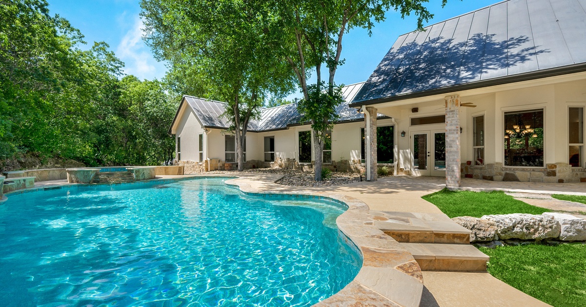 A curved backyard swimming pool with clear blue water next to a white house, stone patio, green lawn, and tall trees.
