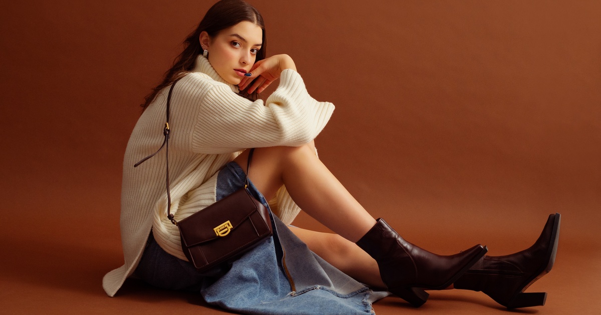 A woman wearing a white sweater, denim skirt, brown ankle boots, and a shoulder bag, standing against a brown background.