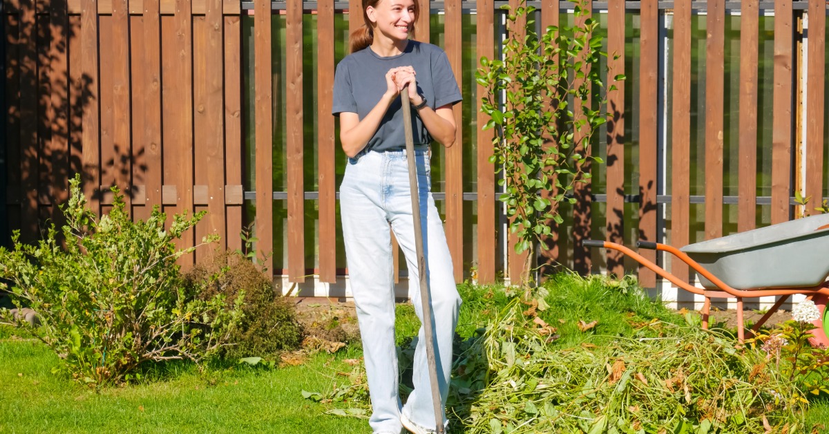 A smiling woman holding a rake beside a pile of garden clippings and a wheelbarrow while standing in a sunny yard.