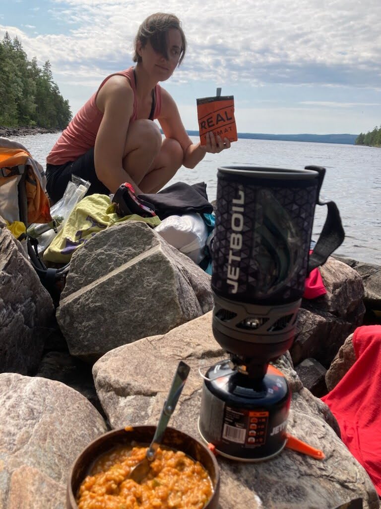 A person by a lake holding a meal bag, with a portable stove and a bowl of food in the foreground.