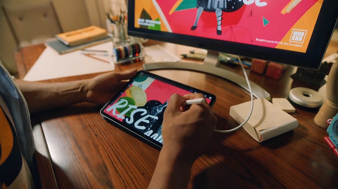 Chico trabajando con un iPad Pro sobre una mesa y enchufado a una pantalla de ordenador. Está usando el Apple Pencil para crear algún tipo de composición publicitaria. Se lee la palabra “RISE” sobre un collage en la pantalla del iPad.
