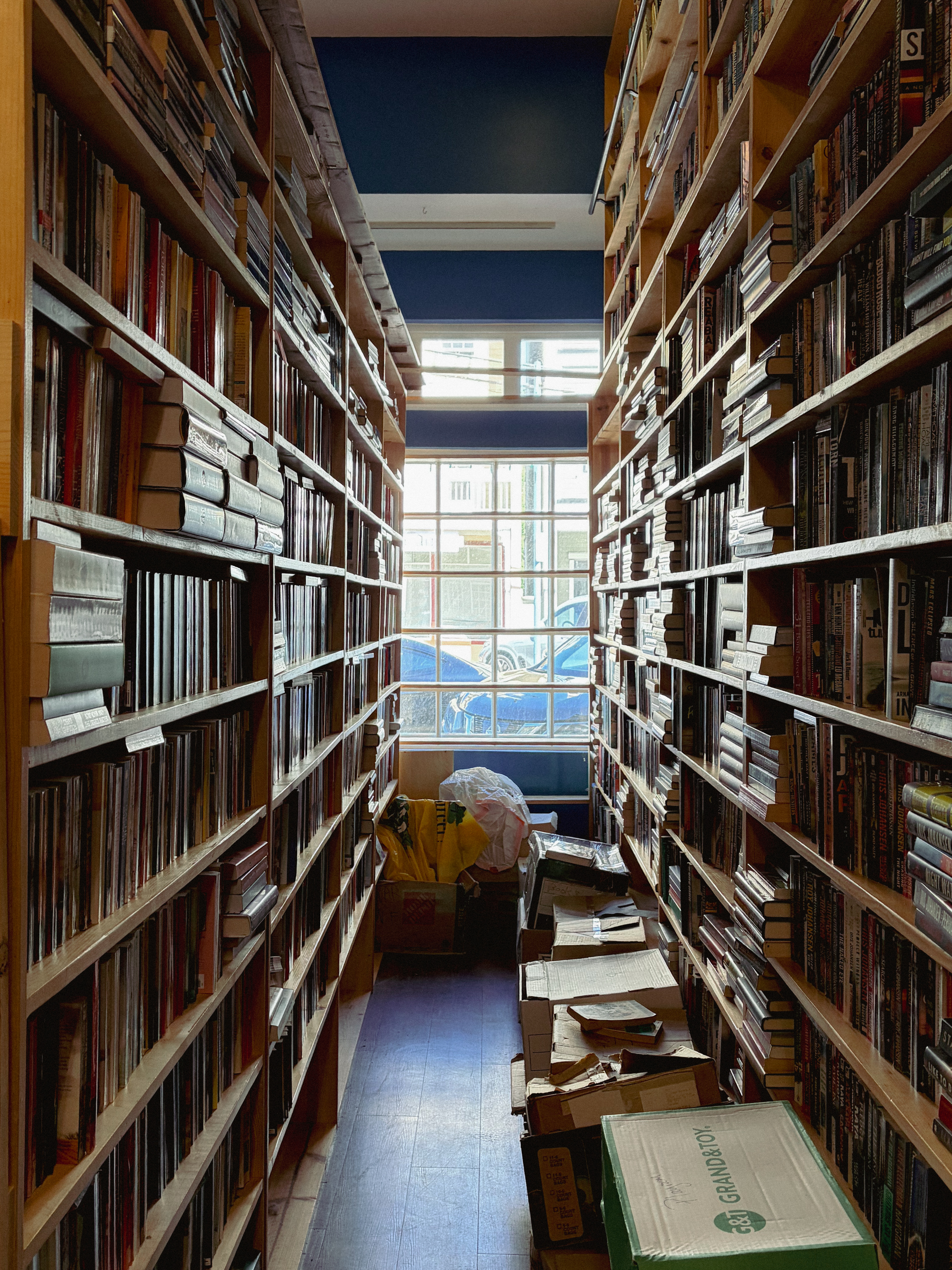 Narrow library aisle between tall book-filled shelves; boxes and bags on the floor near a bright window at the end.