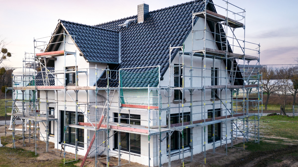 Ein zweistöckiges Einfamilienhaus mit dunklem Ziegeldach entsteht, eingerahmt von einem Gerüst. Die weißen Außenwände deuten auf modernes Design hin, während das Rasengrundstück auf die Landschaftsgestaltung wartet. Mit Smart-Home-Elektroplanung im Hinterkopf verspricht dieses Haus modernsten Komfort unter freiem Himmel.