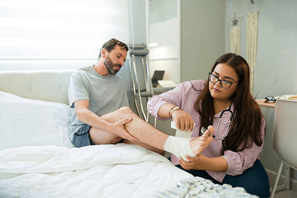 Nurse wrapping bandage around male patient's injured ankle during home visit after surgery, providing post-operative care and support