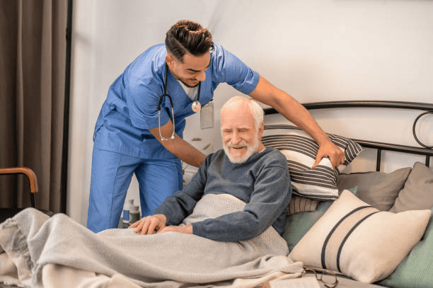 Caring young in-home male nurse putting pillows under the back of a pleased old man