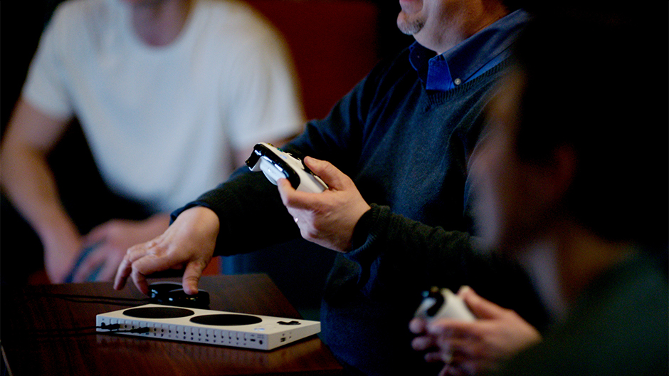 A person uses an Xbox Adaptive Controller on a table while holding an Xbox Wireless Controller