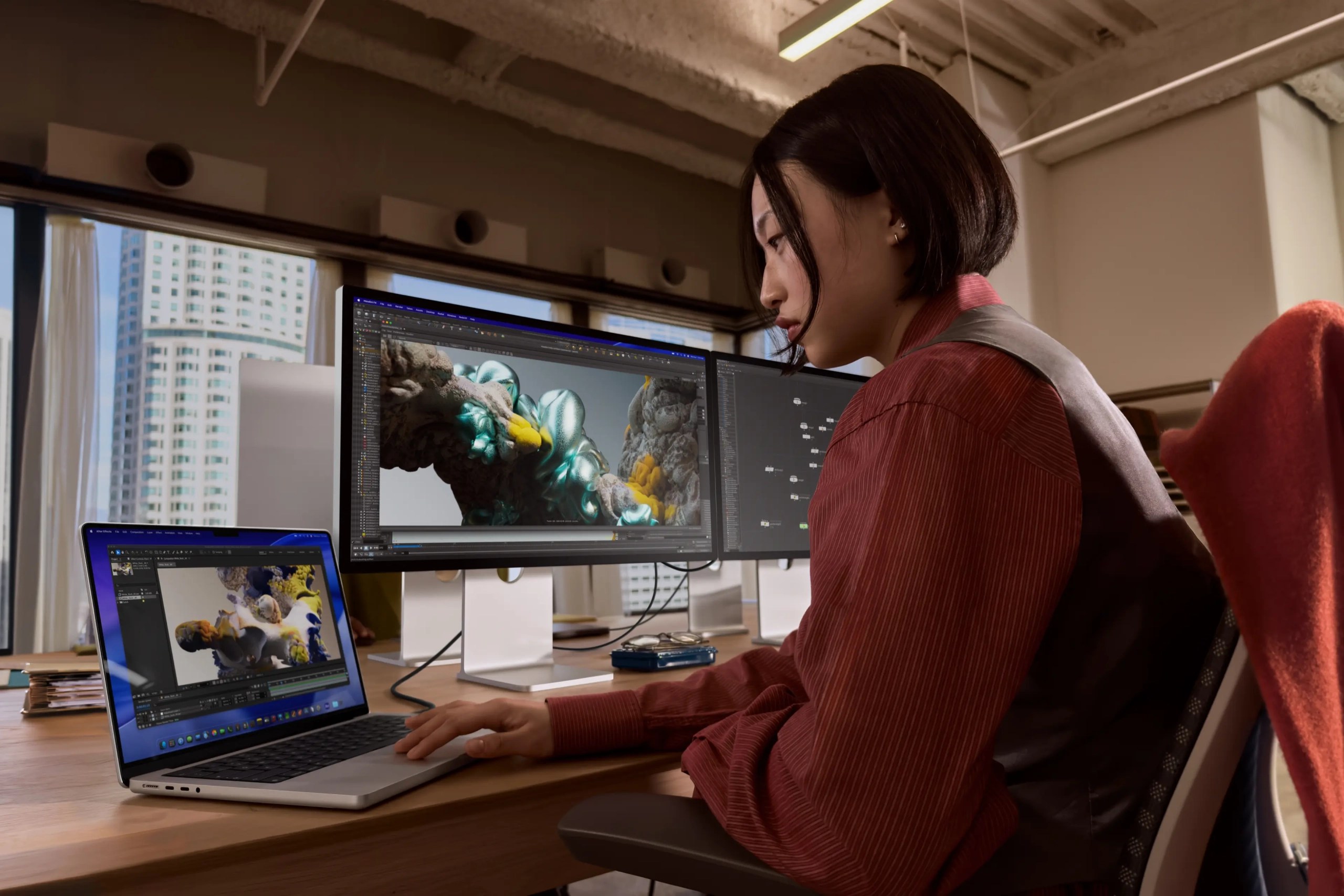 A woman working on a laptop at a modern office desk, with two large monitors displaying intricate 3D graphics. She is focused and wearing a striped shirt, with a cityscape visible through the windows in the background.
