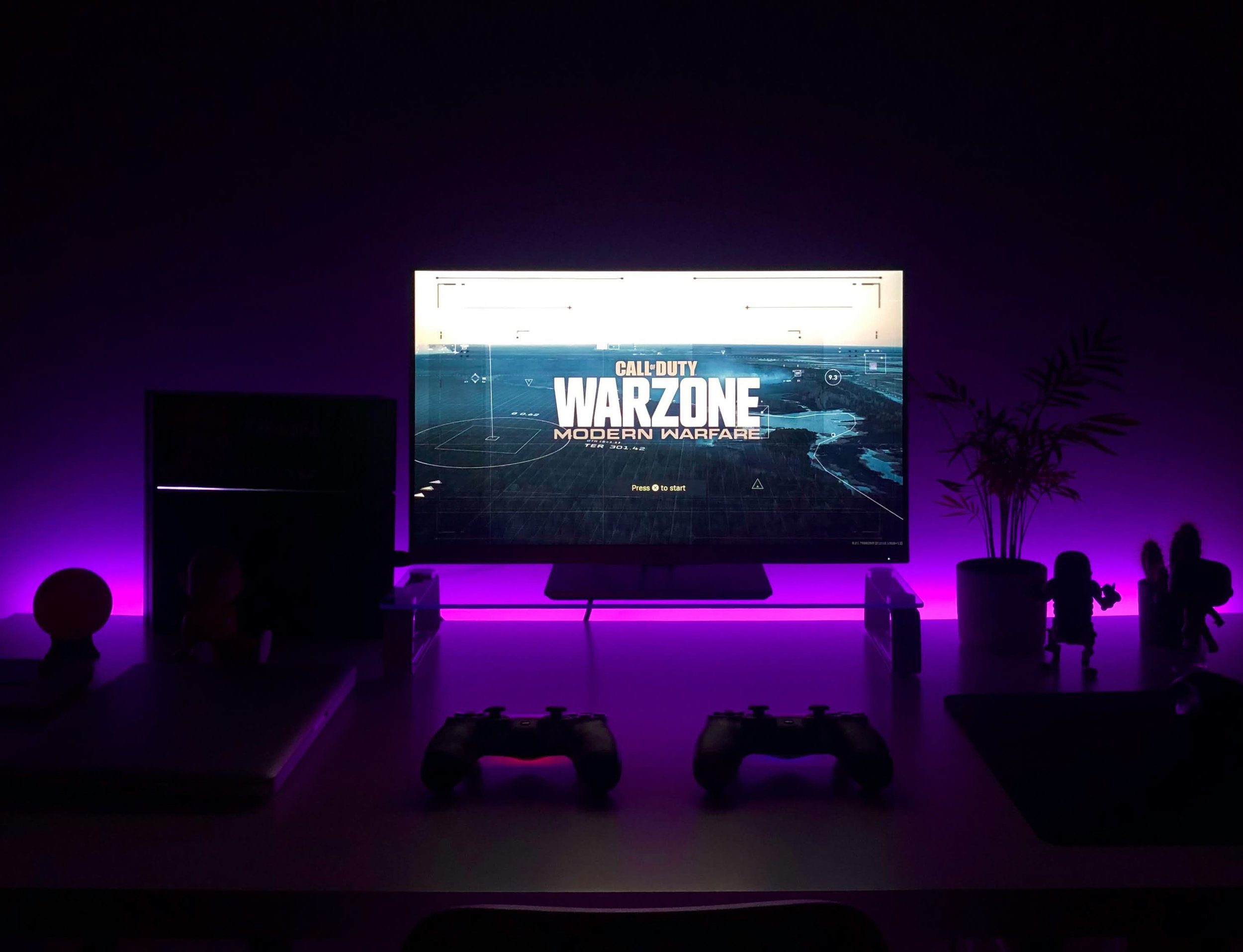 A gaming setup with a monitor displaying Call of Duty: Warzone, flanked by purple LED lights, PlayStation controllers, and desk accessories