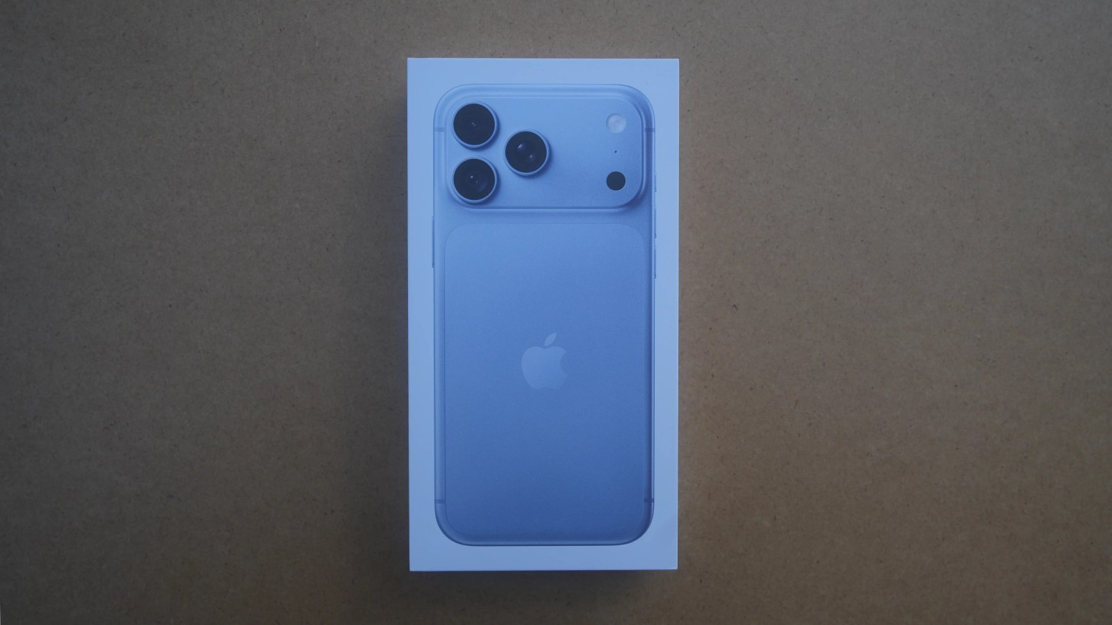An unopened smartphone box featuring a purple iPhone with three rear cameras, displaying the Apple logo on a neutral background.