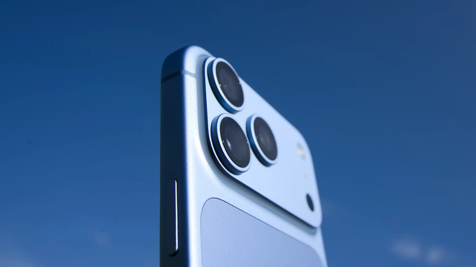 Close-up of a blue smartphone showing its three camera lenses against a clear blue sky.