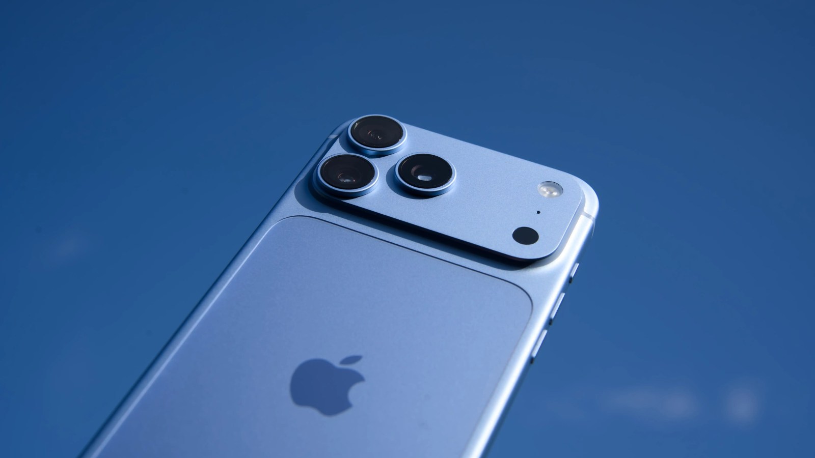 A blue smartphone with a unique triple-camera module is held against a clear blue sky.