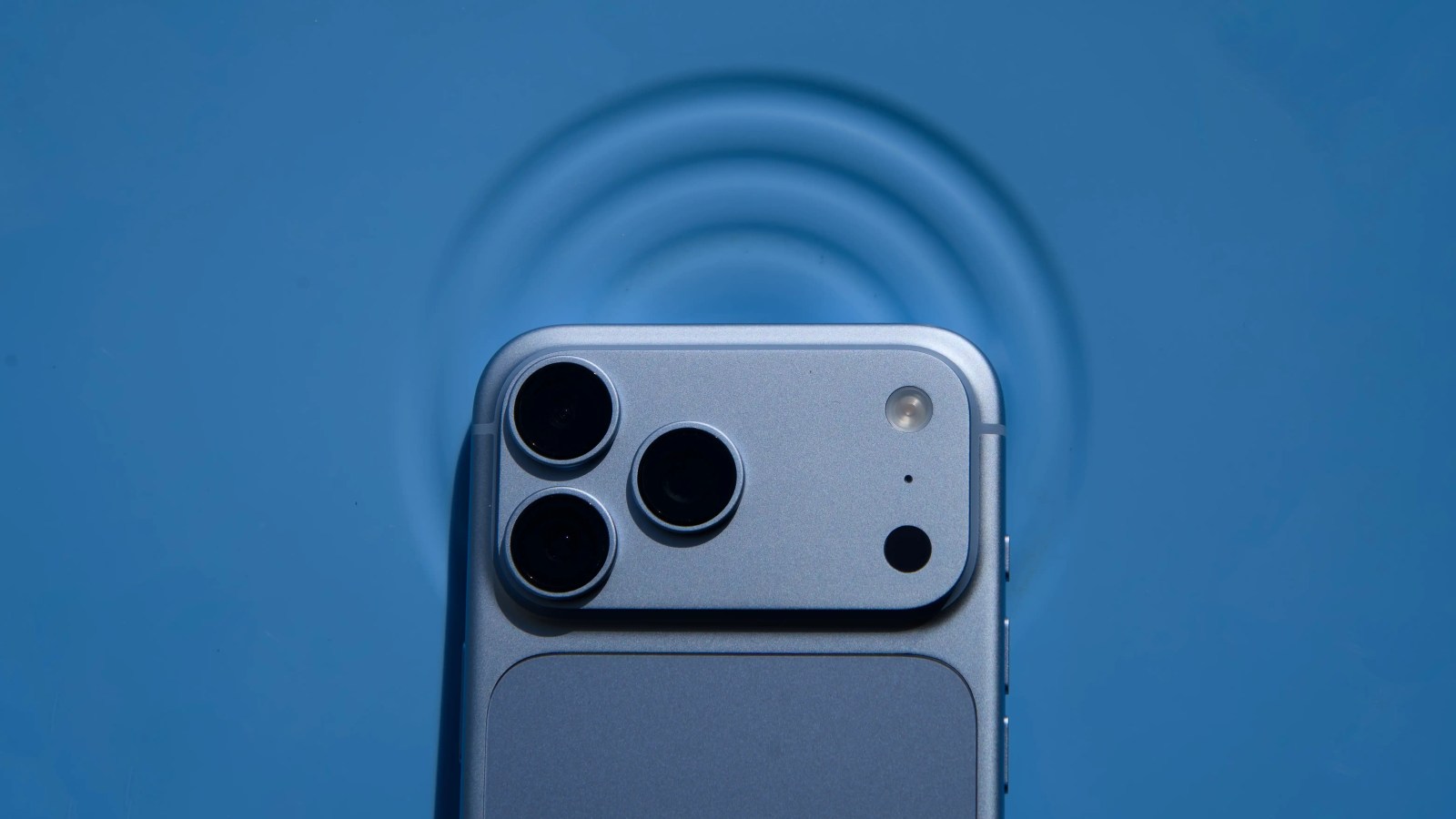 A light blue smartphone with a triple-camera setup lies on a matching blue surface with circular ripples.