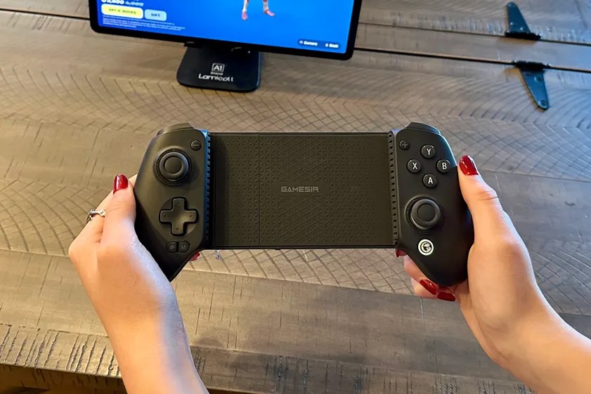 A person with red nail polish holds a black GameSir mobile gaming controller over a wooden table.
