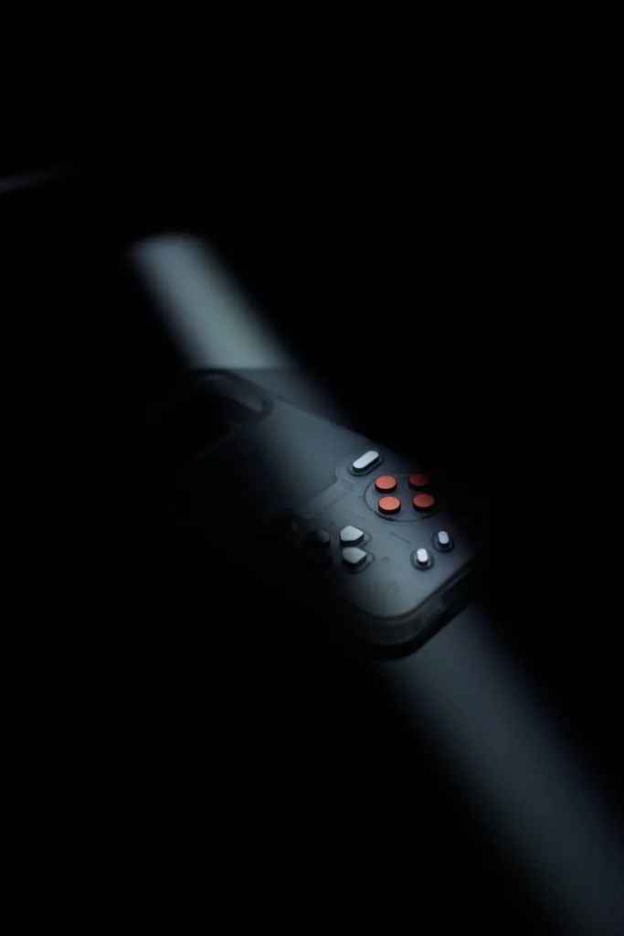 A handheld gaming console with orange buttons is partially illuminated by a dramatic beam of light in darkness.