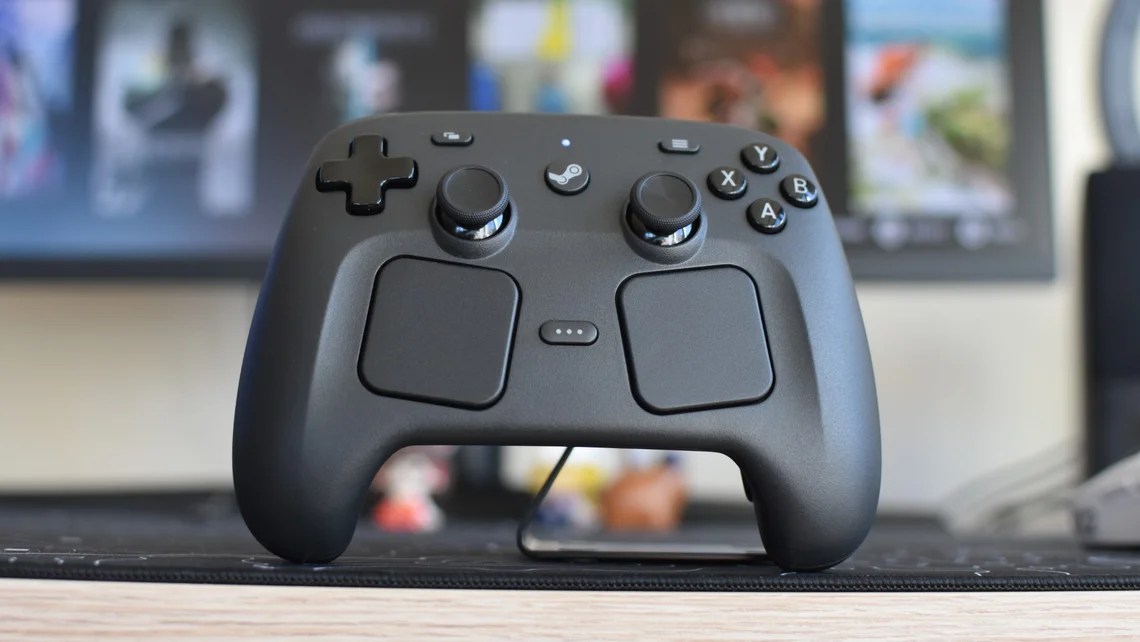 A matte black Steam controller with dual trackpads and joysticks sits on a desk before a blurred monitor.