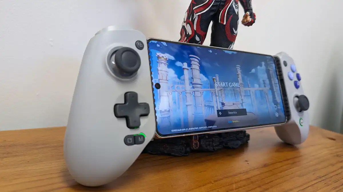 A mobile phone game controller holds a smartphone displaying a video game start screen on a wooden surface.