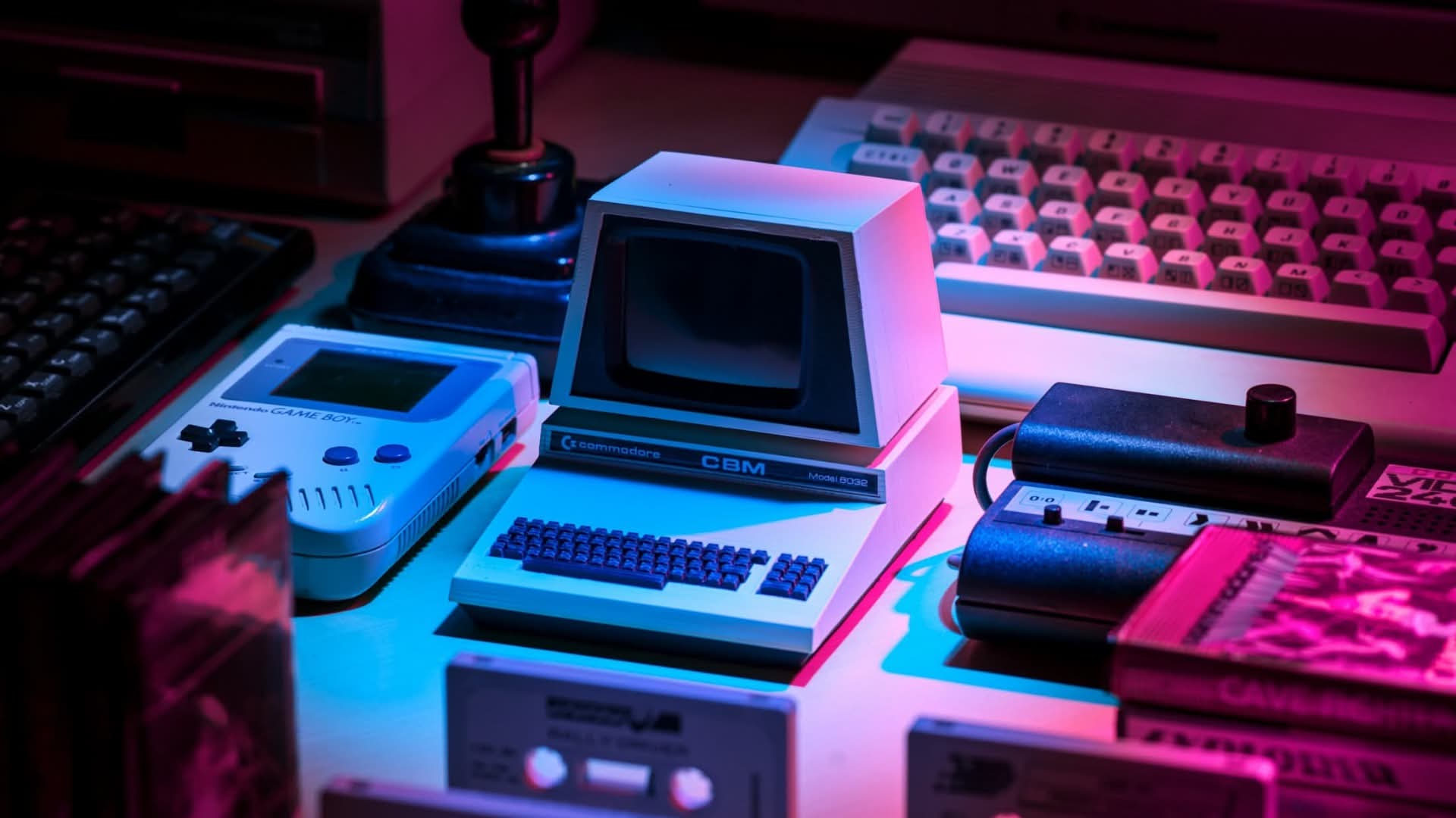 A retro-themed desk displays a vintage computer, a handheld gaming device, an old joystick, and a keyboard. Cassettes, books, and a game controller are also present, all bathed in pink and purple lighting.