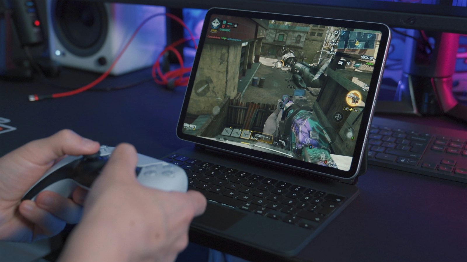 A person playing a video game on a tablet connected to a keyboard, using a game controller. The game displayed is a first-person shooter, featuring an action scene with a character aiming a weapon.