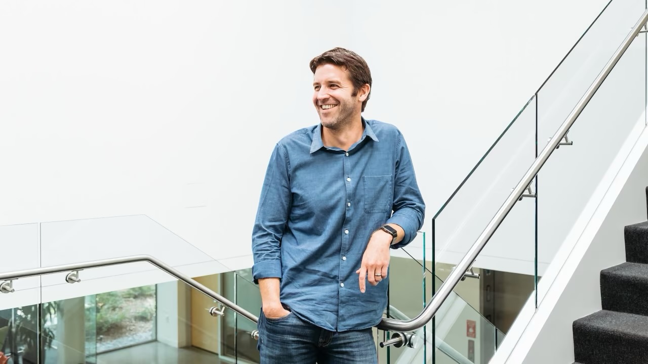 A smiling man in a blue button-down shirt leans against a glass staircase railing in a modern office.