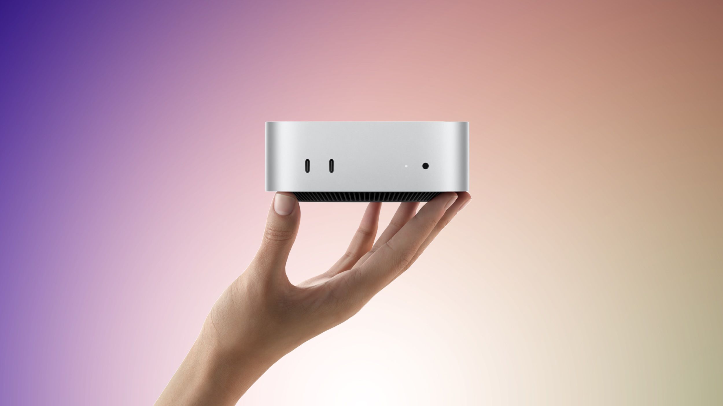 A person's hand gently holds a compact, silver Mac mini desktop computer against a gradient background.