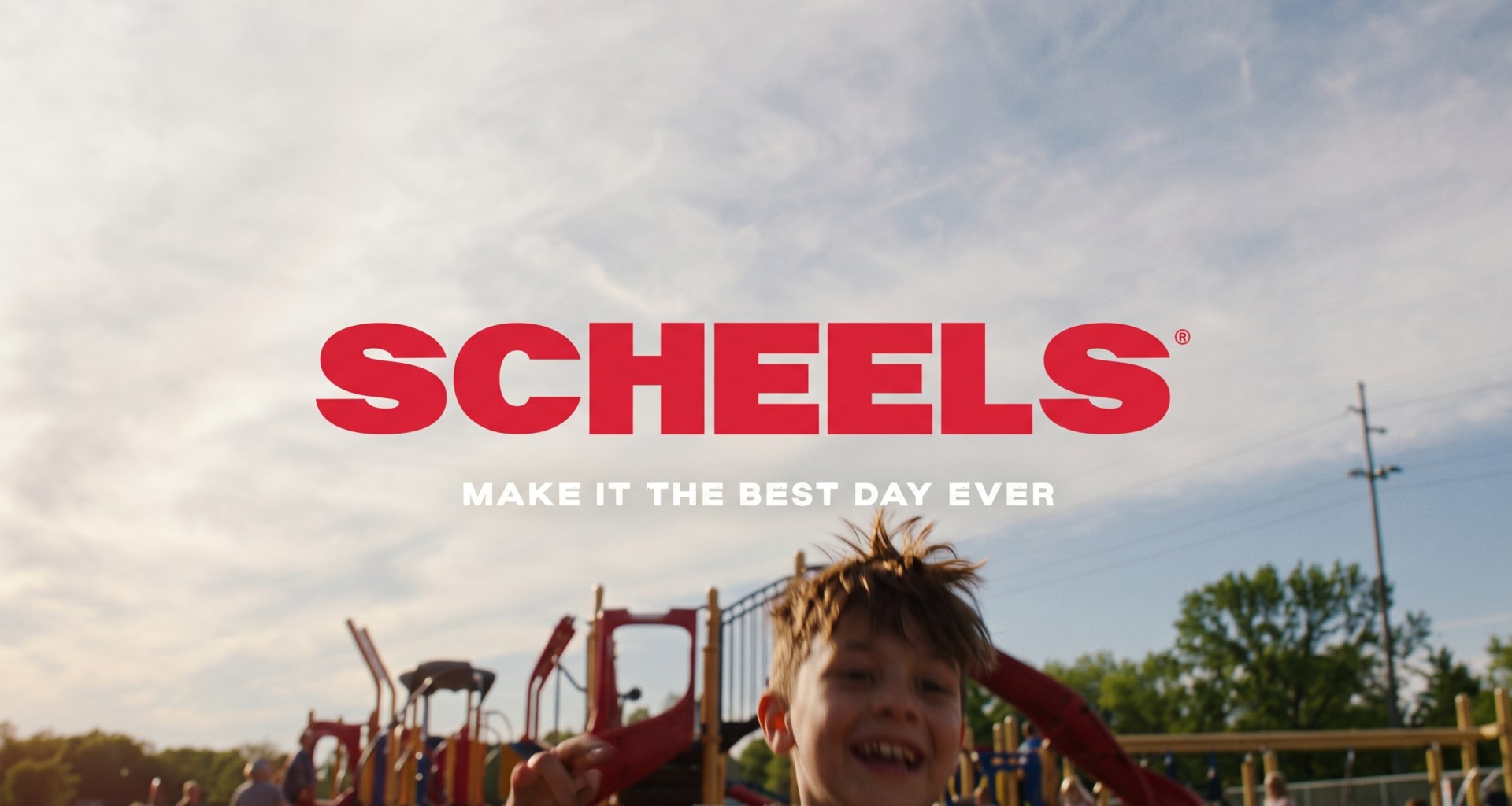 Scheels “Make It The Best Day Ever” brand campaign banner featuring a laughing young boy running on a playground under a bright cloudy sky.