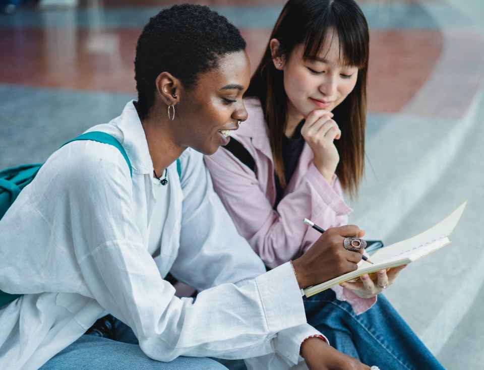 smiling black woman sharing workbook with crop asian girlfriend outdoors