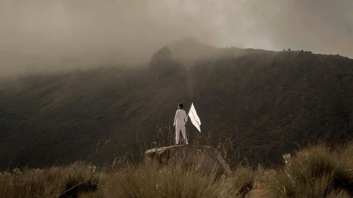 A man in white holding a white flag stands on a large rock surrounded by mountains, symbolizing surrender and courage in faith.