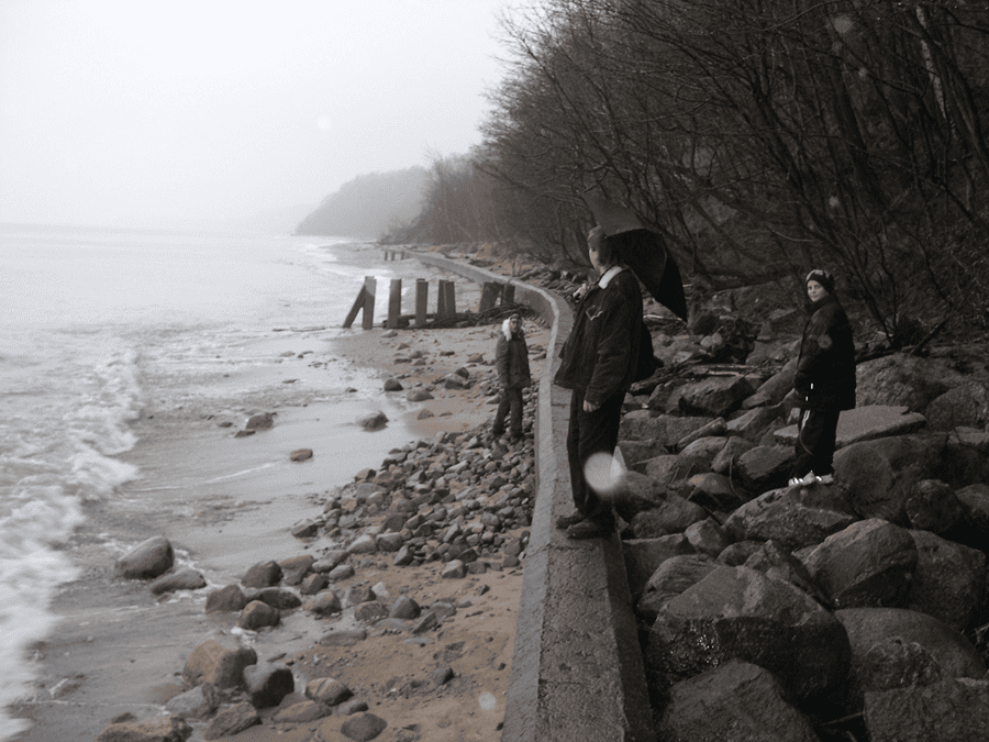 Three people in coats stand on a rocky seawall along a misty, grey beach, one holding a black umbrella as waves wash the shore. Victor Ryabinin. Kaliningrad artist, leads this excursion