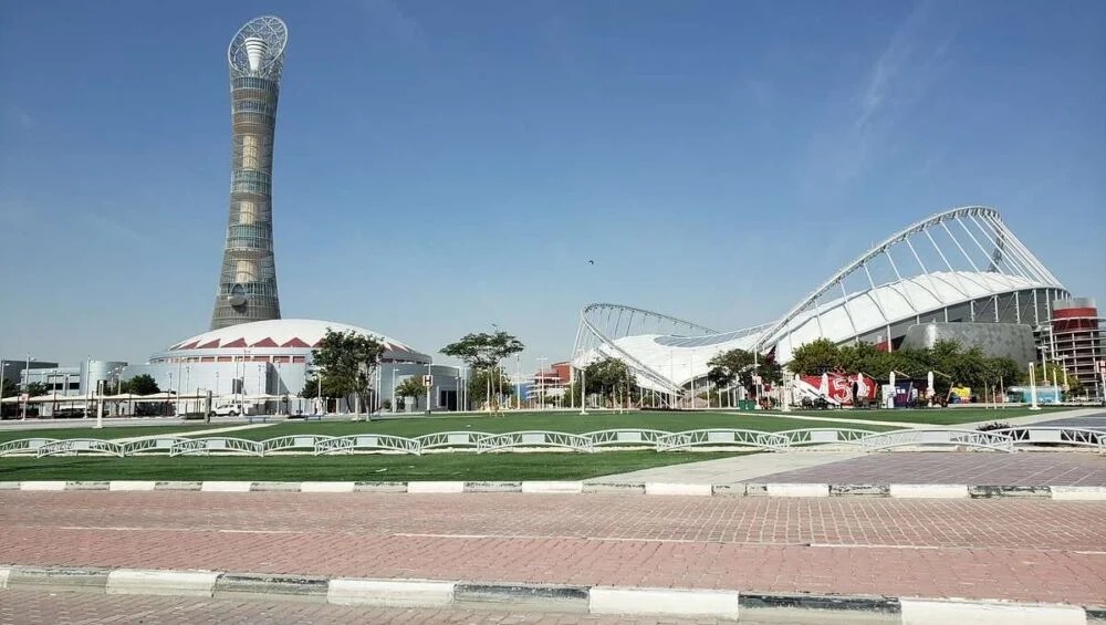A tall, spiral-shaped tower stands beside a stadium with a curved roof under a clear blue sky, with green lawns and trees—an ideal setting for Qatar National Sports Day—while people stroll along the paved walkway in the foreground.