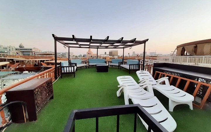 Rooftop seating area with artificial grass, white lounge chairs, blue cushioned seats, and a wooden pergola, overlooking a cityscape at sunset—perfect for unwinding after a Traditional Dhow Boat Rental experience.