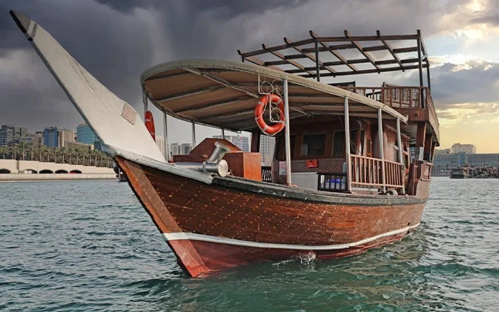 A traditional dhow boat rental floats on calm water under a cloudy sky, with city buildings and palm trees visible in the background.