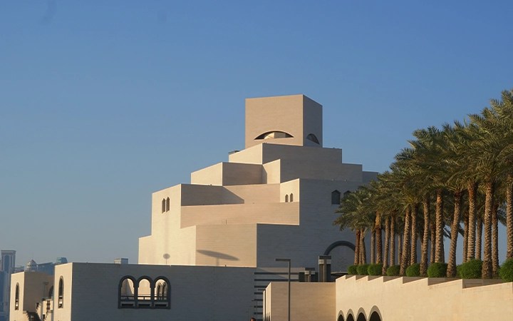 A modern, geometric building with stacked, cream-colored blocks stands beside a row of palm trees under a clear blue sky—its sharp lines and arched windows echo the striking landscapes seen on a half day desert safari Qatar.