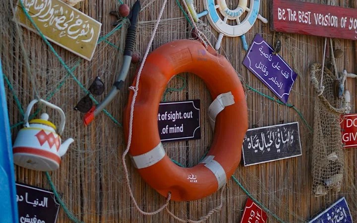 An orange lifebuoy with silver tape hangs on a wall covered in fishnet, colorful signs in Arabic and English, a teapot, sunglasses, and a steering wheel—creating the perfect backdrop for your Full Moon Kayaking Qatar adventure.