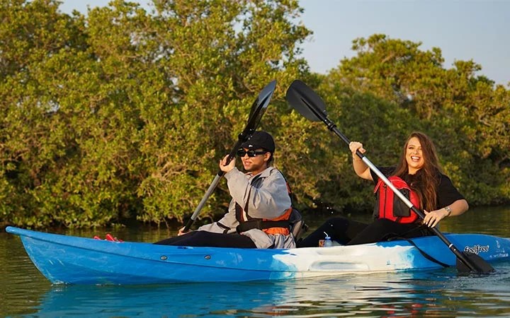 Two people, a man and a woman, paddle a blue kayak together on calm water under the glow of Full Moon Kayaking Qatar, surrounded by green trees and sunlight. Both wear life jackets and appear to be enjoying themselves.