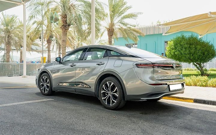 A silver Toyota Camry sedan parked on a street in Qatar, framed by palm trees and a turquoise building in the background on a sunny day.