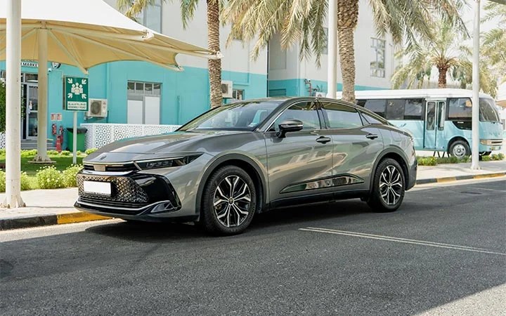 A sleek, silver Toyota Camry Qatar is parked on a city street lined with palm trees. A blue and white bus and a turquoise building with large windows are visible in the background.