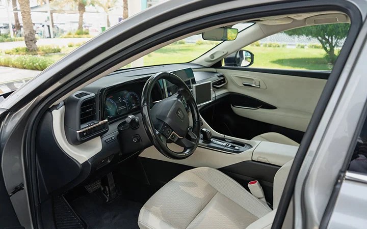 The interior of a modern Toyota Camry Qatar features a black steering wheel, digital dashboard, touchscreen display, and light-colored fabric seats, all viewed from the open driver’s side door.