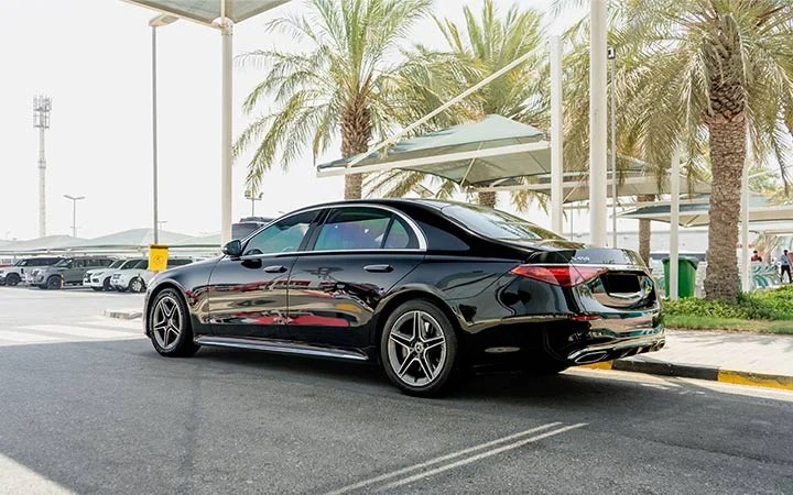 A sleek black Mercedes S Class Qatar luxury sedan is parked on the roadside under a canopy, with palm trees and several other vehicles visible in the background on a sunny day.