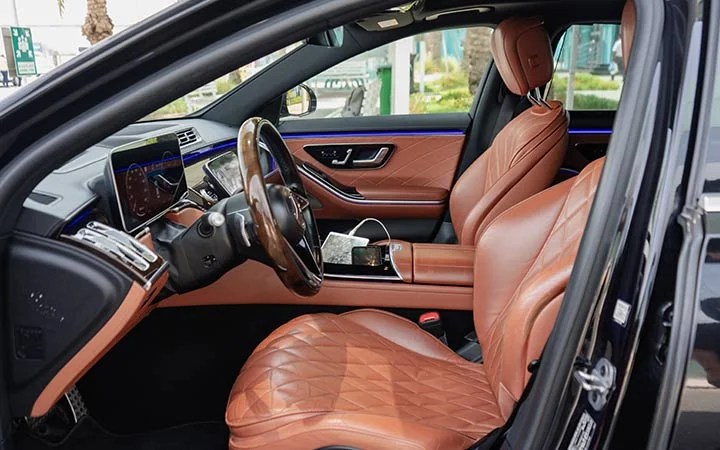 The image shows the luxurious interior of a Mercedes S Class Qatar with tan diamond-stitched leather seats, a digital dashboard, and a smartphone connected by a charging cable on the center console.