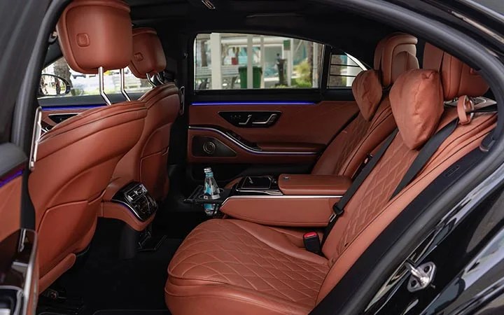 Luxury car interior with brown diamond-stitched leather seats, ample legroom, armrest controls, a bottle of water on the console, and ambient blue lighting—experience true comfort in a Mercedes S Class Qatar.